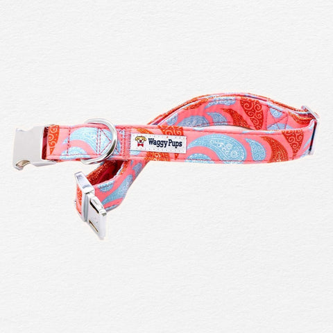 The image shows a dog collar with a red and blue paisley pattern. The collar is made of a durable fabric material and has a silver buckle on one end. The buckle is attached to the collar with two metal clips. On the collar, there is a small white label with the brand name "Waggy Pups" written in black. The background is plain white.
