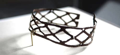 The image shows a close-up of a dog collar on a white surface. The dog collar appears to be made of metal and has a unique design with a lattice-like pattern. The lattice pattern is made up of small, interlocking lines that create a geometric shape. The metal is a dark brown color and is slightly tarnished, indicating that it has been exposed to the elements for some time. There is a gold-colored metal hook attached to the top of the lattice, which is likely used to attach a leash. The background is blurred, so the focus is on the collar.