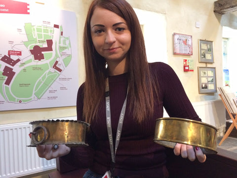 The image shows a young woman standing in a room with a map of a building on the wall behind her. She is holding two brass dog collars in her hands and is smiling at the camera. The woman is wearing a maroon sweater and has a lanyard around her neck. She appears to be in a museum or exhibition space with other exhibits and posters on the walls. The map on the left side of the image shows the layout of the building and the location of the museum.