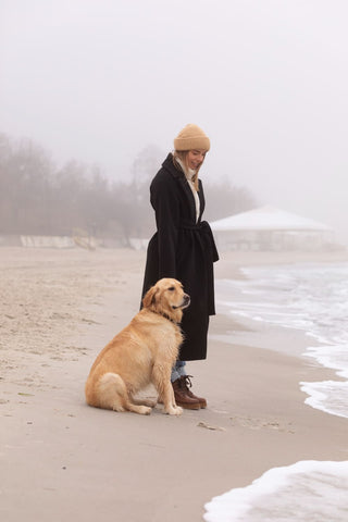 The image shows a woman standing on a sandy beach with a golden retriever dog. The woman is wearing a black coat and a beige beanie, and she is looking down at the dog with a smile on her face. The dog is sitting on the beach next to her, and the ocean can be seen on the right side of the image. The sky is overcast and foggy, and there are trees in the background.