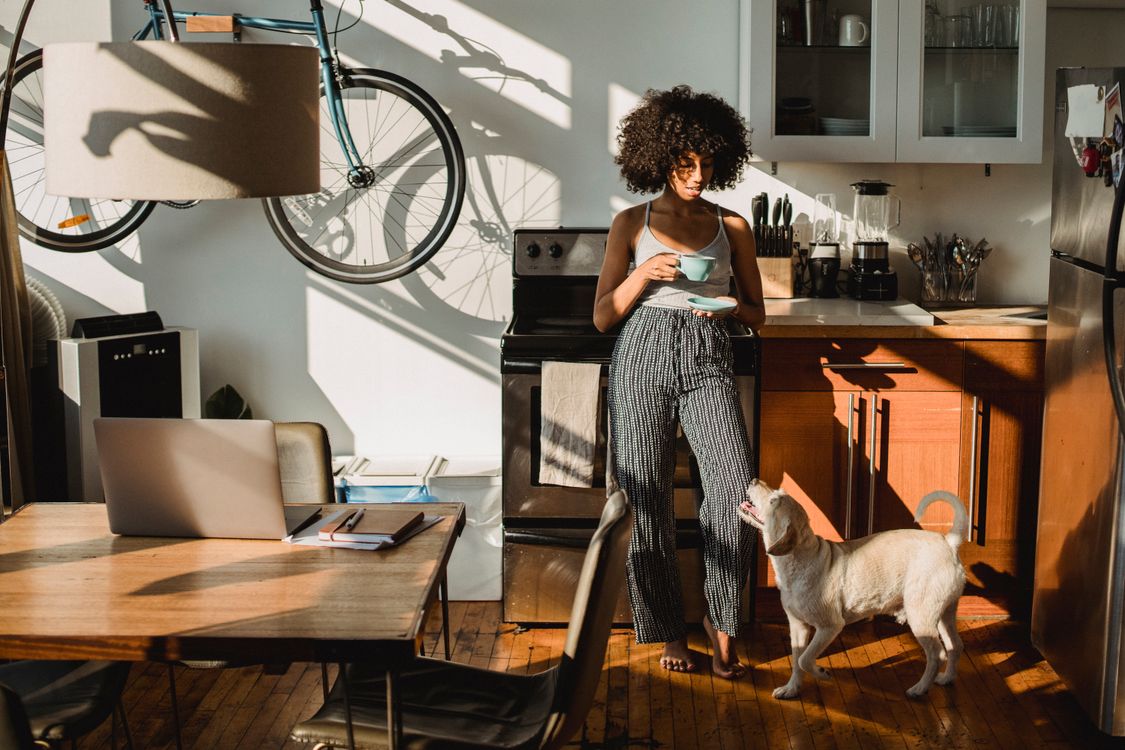 woman drinking coffee in a kitchen while her dog sits at her feet
