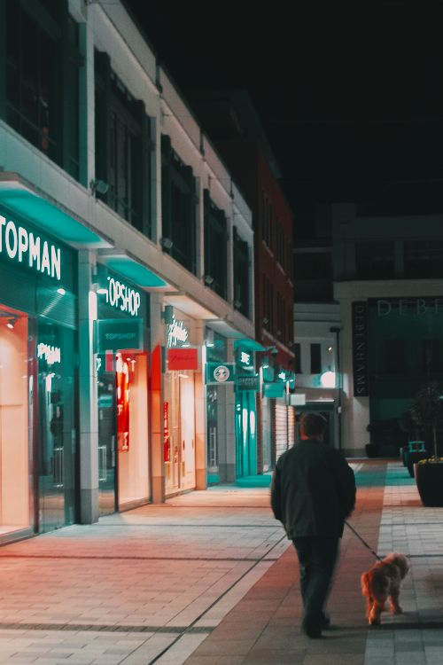 A person walking a dog along a brightly lit street at night